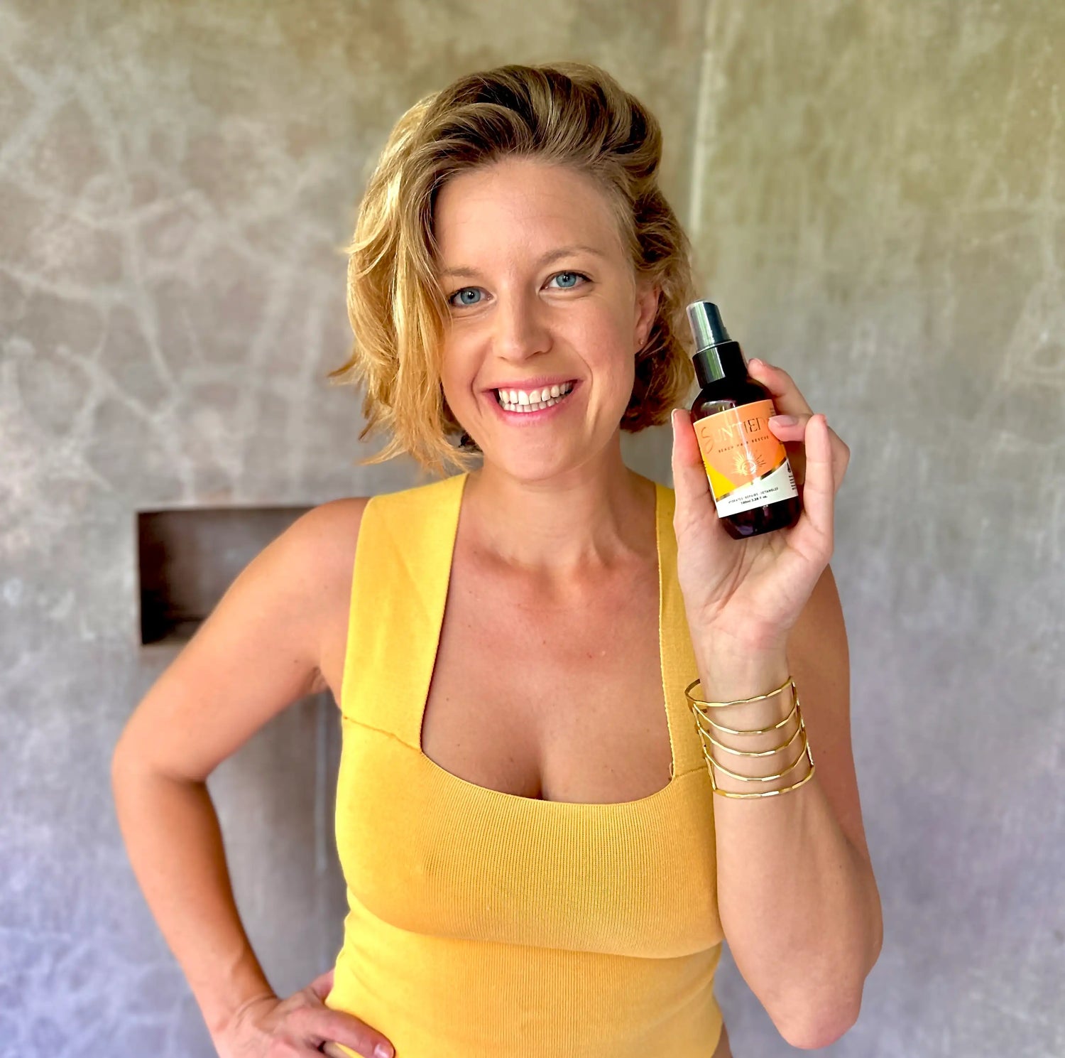 Blonde Woman in yellow top, holding a bottle of Suntied Leave-in conditioner spray 